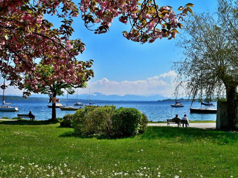 Menschen sitzen am Starnberger See auf Bänken und schauen aufs Wasser mit den Alpen am Horizont.